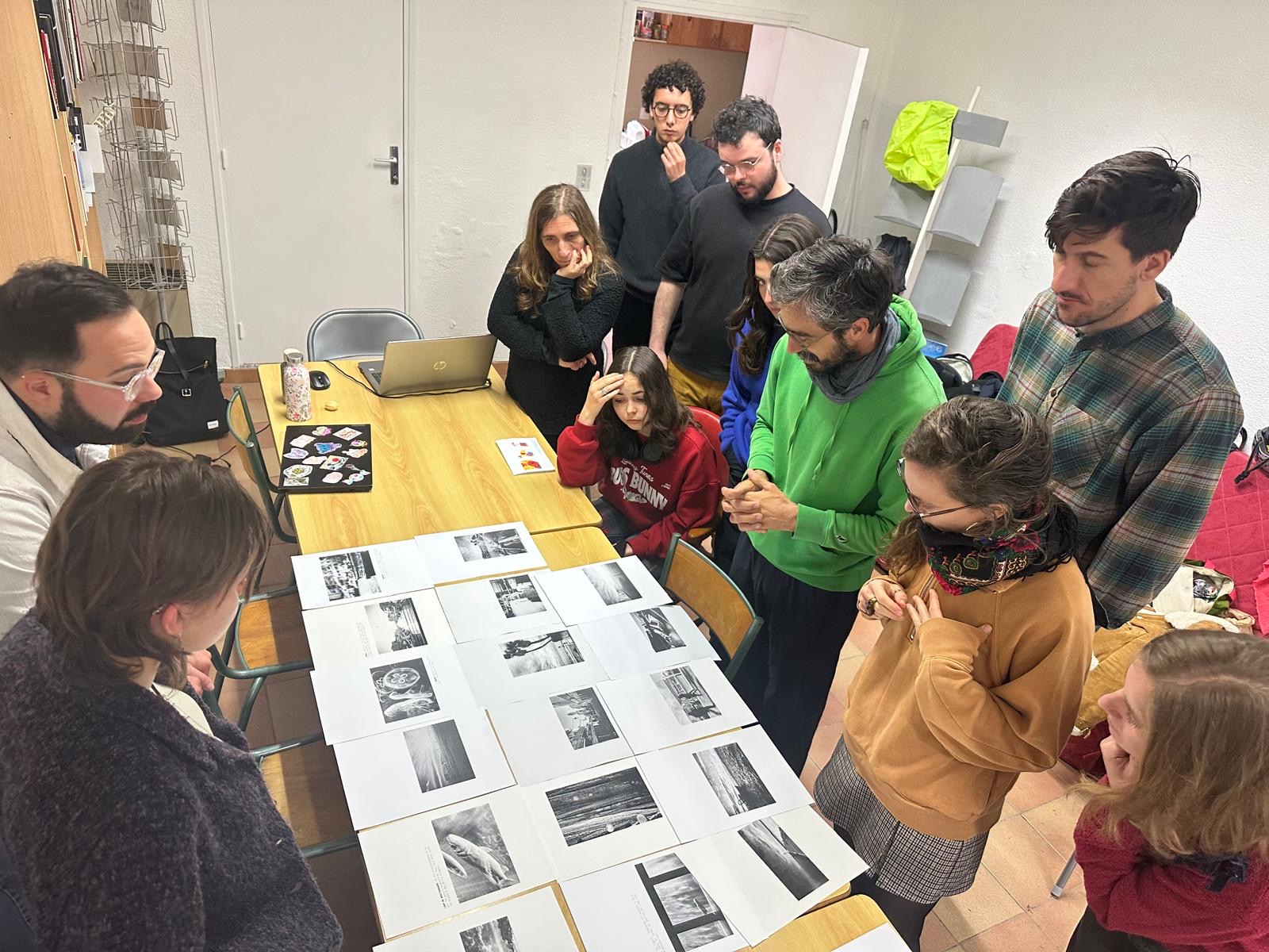 Andrea Olga Mantovani lors d'un workshop de photographie, entourée de stagiaires examinant des tirages photographiques posés sur une table. L'ambiance est studieuse et créative.
